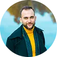Smiling man with short hair and beard wearing a yellow sweater and dark jacket outdoors.