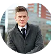 Young man in a dark coat and tie standing outdoors with blurred buildings in the background.