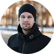 Man wearing a black beanie and black jacket stands outdoors with city lights blurred in the background.