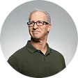 Mature man with gray hair and glasses wearing a dark green polo shirt against a gray background.